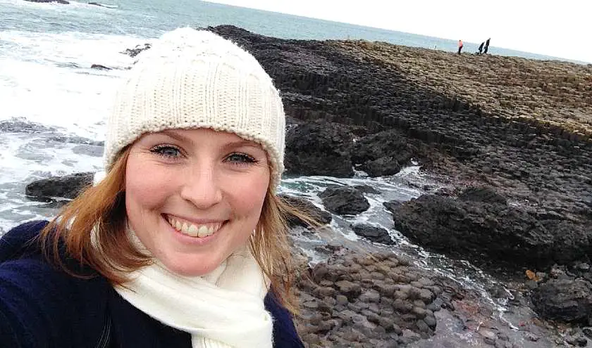 Mel taking a selfie wearing a white wooly hat in front of the Giant's Causeway with the waves crashing in the background