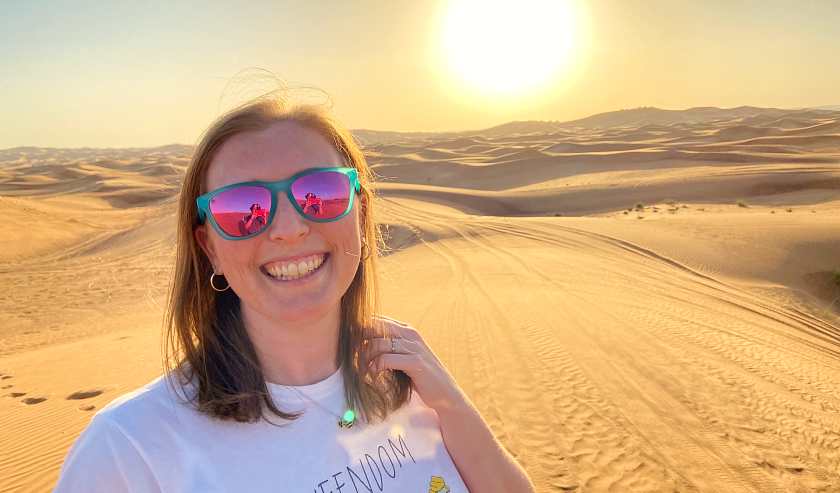 Mel smiling in the Dubai desert wearing pink and green sunglasses and a white t-shirt 