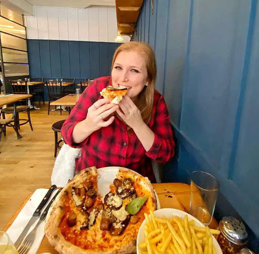 Mel eating a pizza slice in a pizzeria in Brighton wearing a red chequered shirt