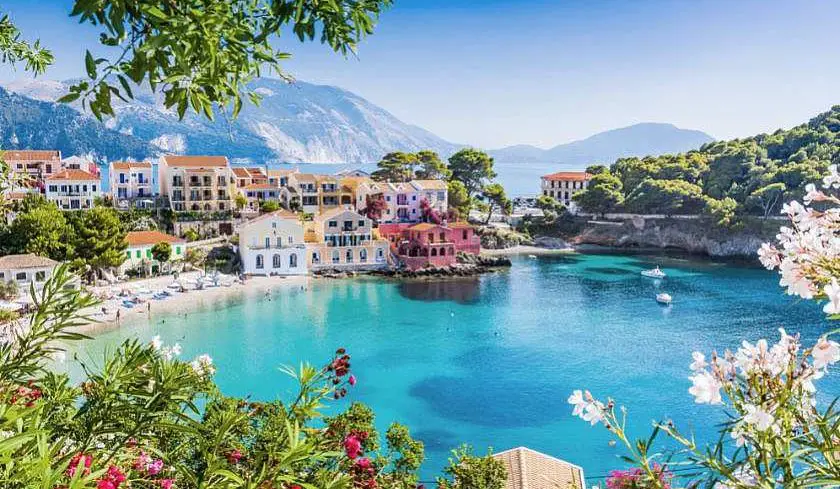 Assos village in Kefalonia with colourful villas around a beach cove and blue water and mountains in the background with flowers and trees framing the photo