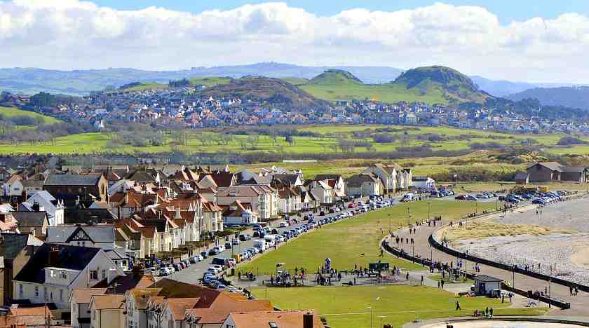 Picture of Llandudno West Shore Beach taken from a far with rows of houses, cars and green hills in the background 