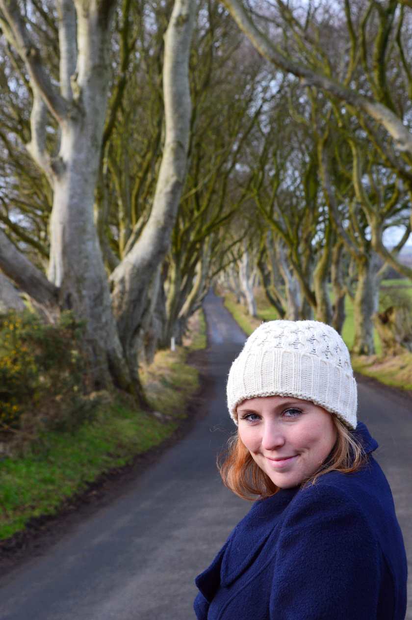 Mel smiling in front of the Dark Hedges wearing a white wooly hat