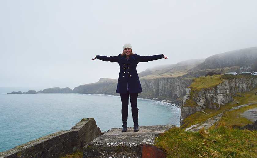 Mel holding out her arms stood at the edge of the coastal waters of Moyle at Limestone Quarry wearing a white hat and blue coat with gold buttons 