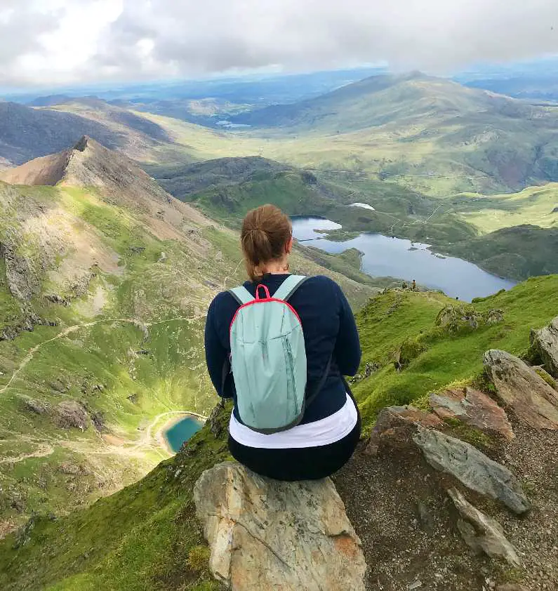 Mel with her back to the camera, sat on a rock with a green backpack on with green peaks and lakes at the summit of Snowdon