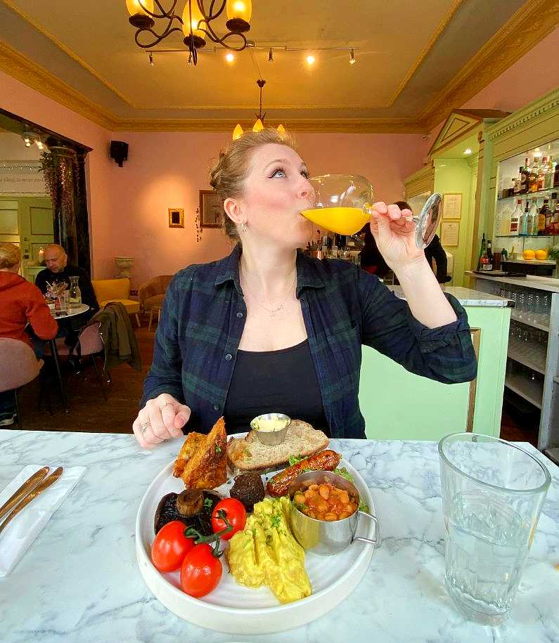 Mel looking up whilst tipping a wine glass of orange juice and champagne into her mouth with a vegan full english breakfast in front of her at Oeuf cafe in Brighton