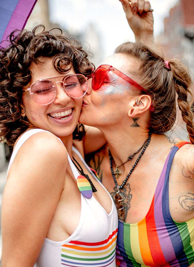 Girl kissing another smiling girl on the cheek both wearing rainbow tops celebrating pride