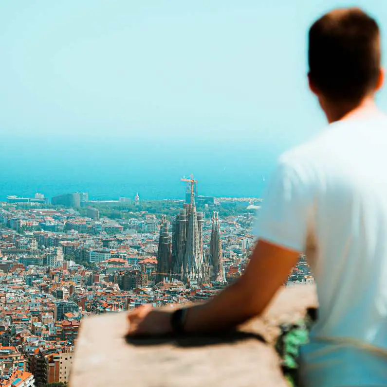 A man wearing a white tshirt looking over a balcony at the Sagrada Familia from a high view point