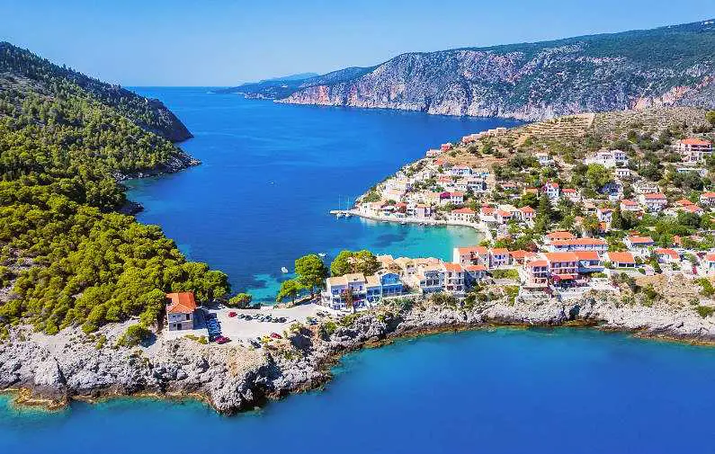 Aerial view of Assos village in Kefalonia with colourful villas around a beach cove and blue water and mountains in the background by the open sea