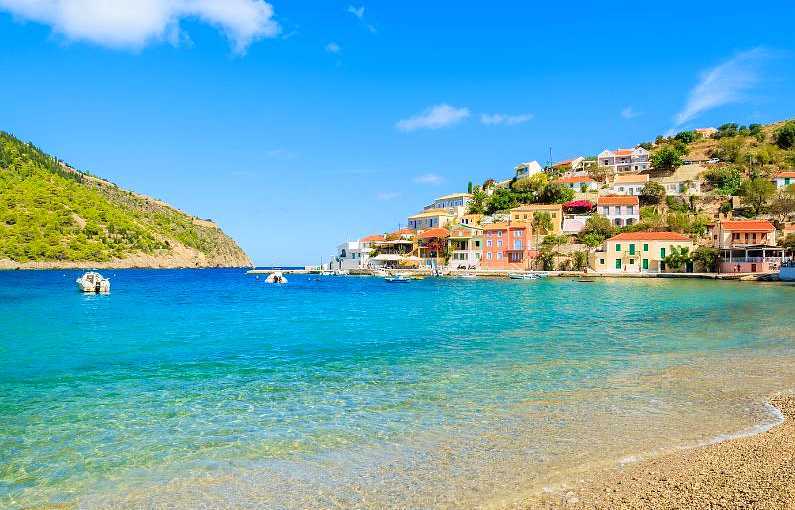 Assos village in Kefalonia with colourful villas around a beach cove and blue water lapping on a sandy beach