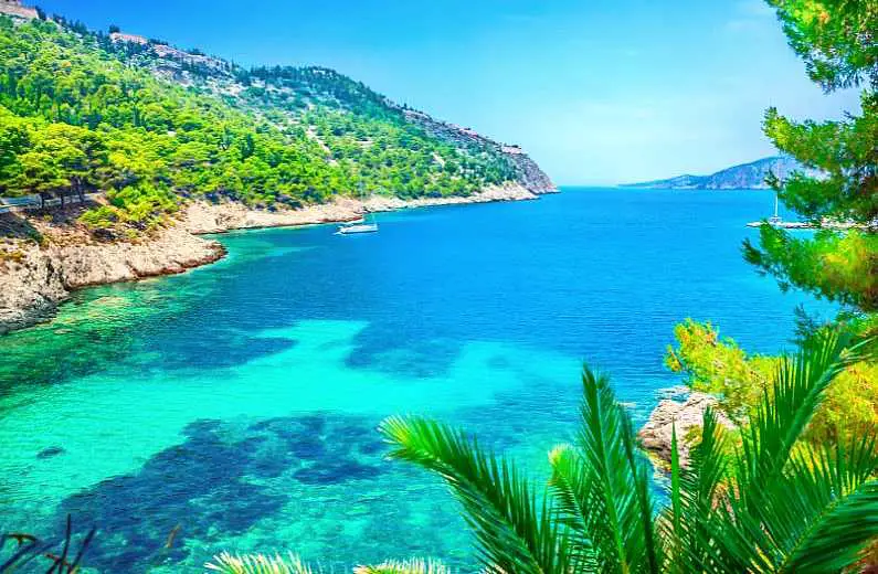 Bright blue and turquoise water in Assos village in Kefalonia with white low cliffs and green trees in the background