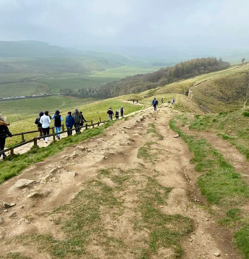 Muddy path to the summit of Mam Tor with people walking down the stone path on the left and the green peaks in the background