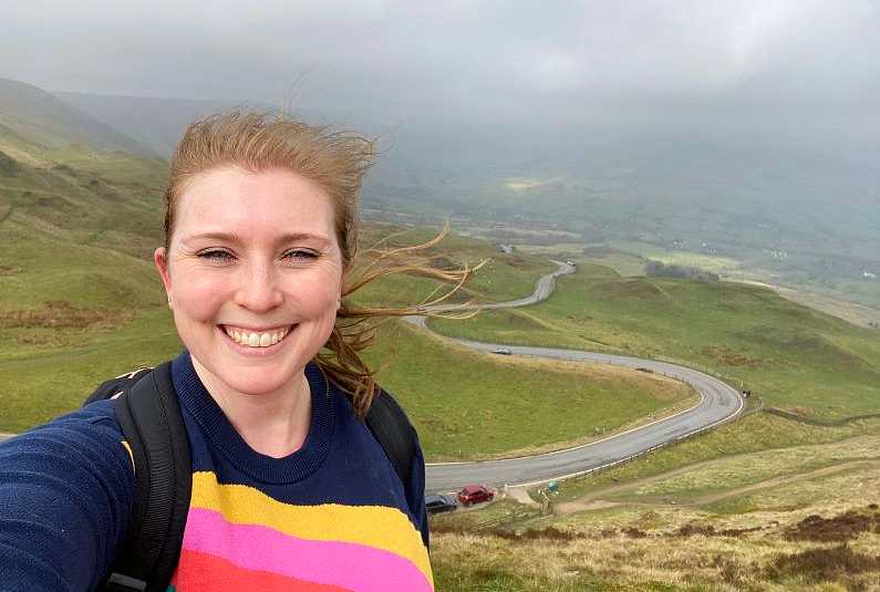 Mel taking a selfie from the top of Mam Tor with wind swept hair and green peaks in the background and a winding road