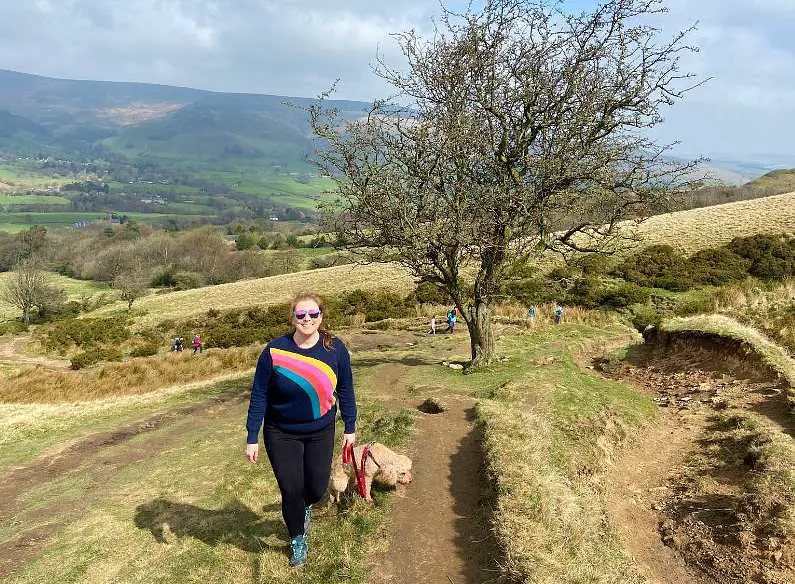 Mel climbing up the muddy path to the top of Mam Tor with her dog Lilly with the green fields in the background and a leafless tree