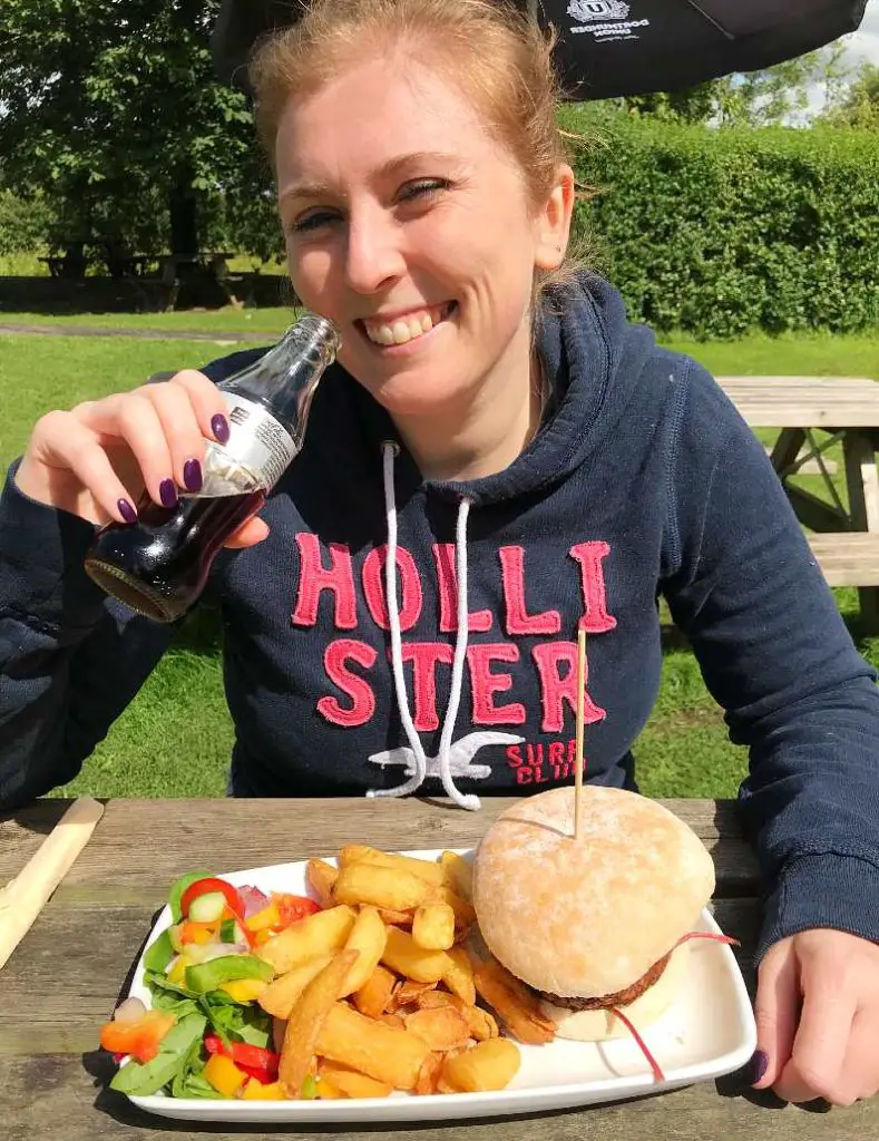 Mel drinking a glass bottle of diet coke and smiling with a vegan burger and chips in front of her in the Ramblers Inn pub in Edale