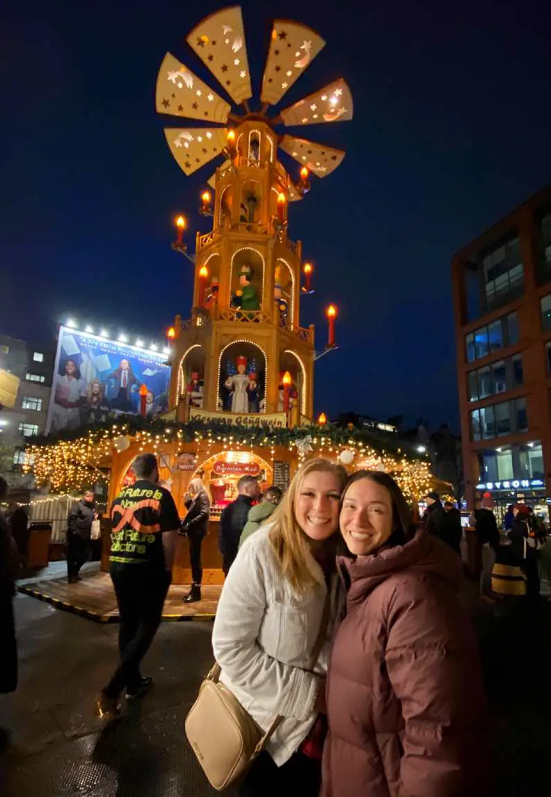 Mel and Vicky smiling at the Christmas Markets in Manchester with a German windmill in the background