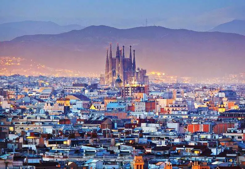 Sagrada familia from a distance at dusk with mountains in the background and small buildings and houses surrounding it 