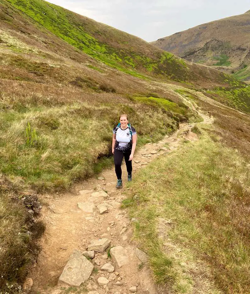 Mel hiking up a dirt path with large peaks behind her in Glossop