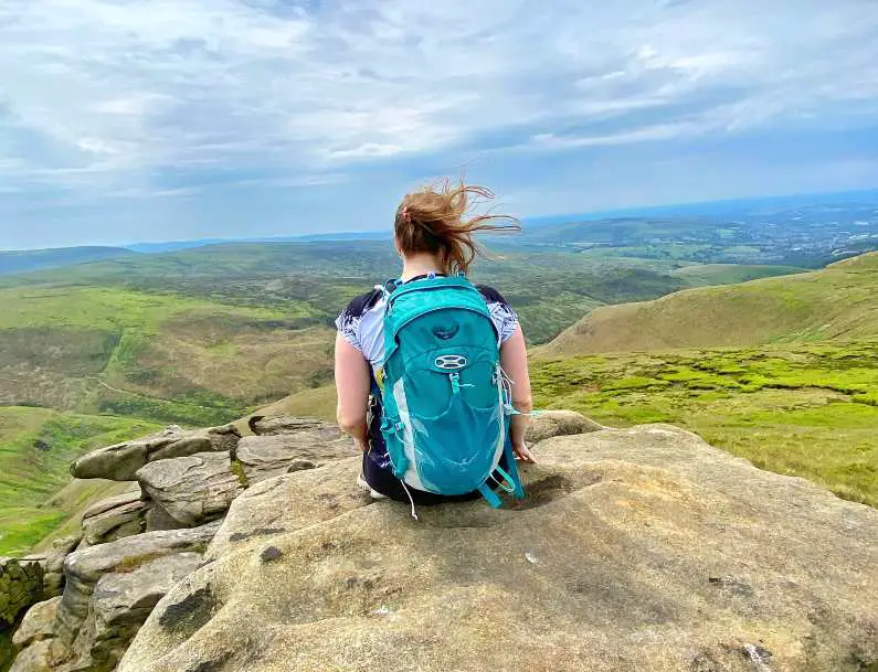 Mel sat on a rock with her back to the camera wearing a green backpack on Bleaklow with the peaks of Glossop in front of her