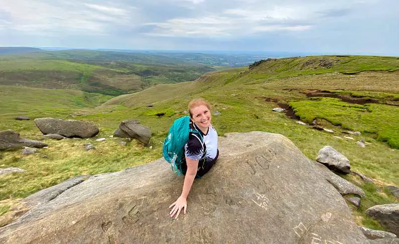 Mel smiling whilst sitting on a rock with the green peaks of Glossop behind her