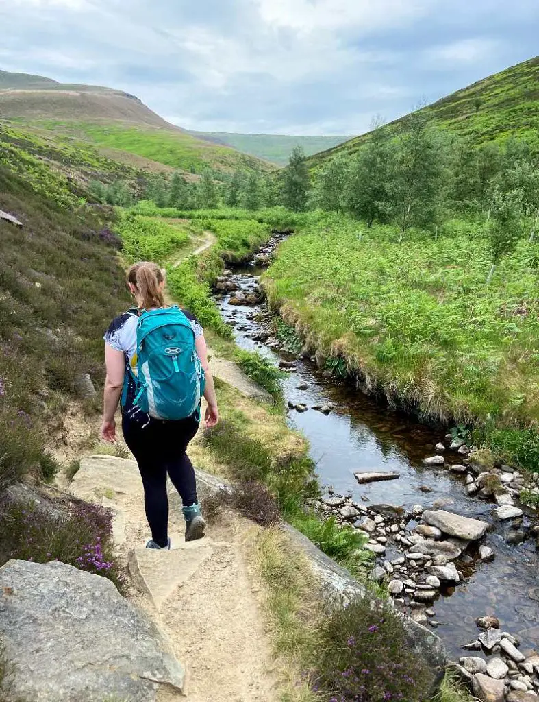 Mel hiking along a stream in the peak district wearing a green backpack and white and black t-shirt