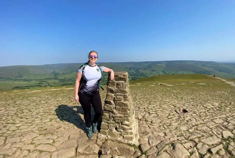 Mel smiling and leaning against the keystone at the top of the Mam Tor summit in the Peak District on a sunny day