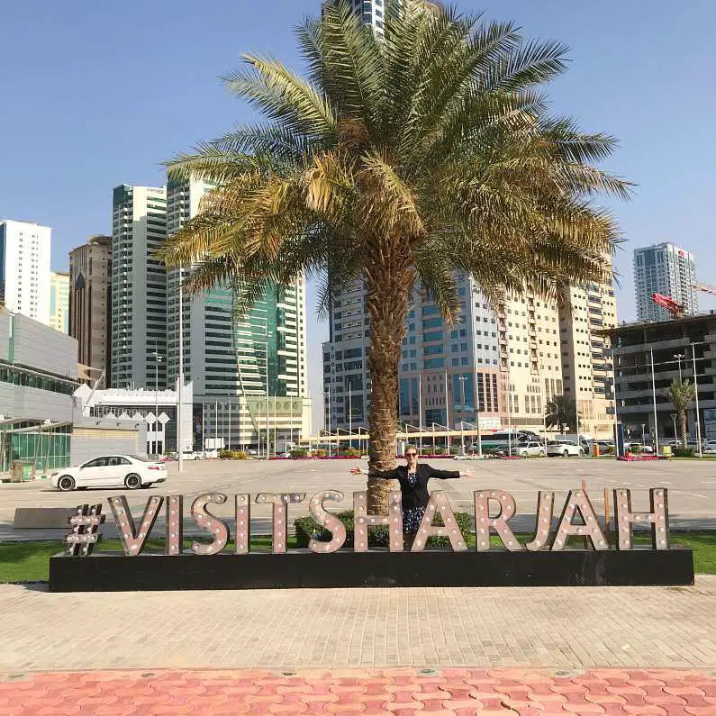 Mel with her arms out, smiling in front of the #VisitSharjah sign in Sharjah, UAE with a palm tree in the background