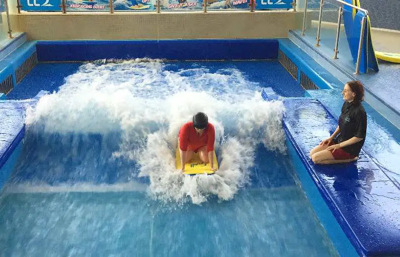 Mel indoor surfing at LC Swansea in an orange swim shirt and a black helmet with an instructor crouched down next to the wave pool 