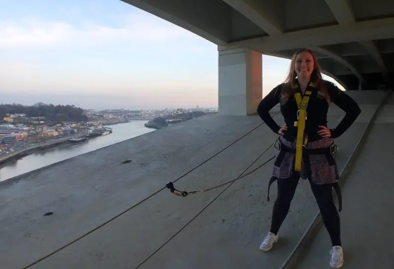 Mel standing in a superman pose in climbing gear, hiking to the top of the Porto Bridge with the city in the background at sunset