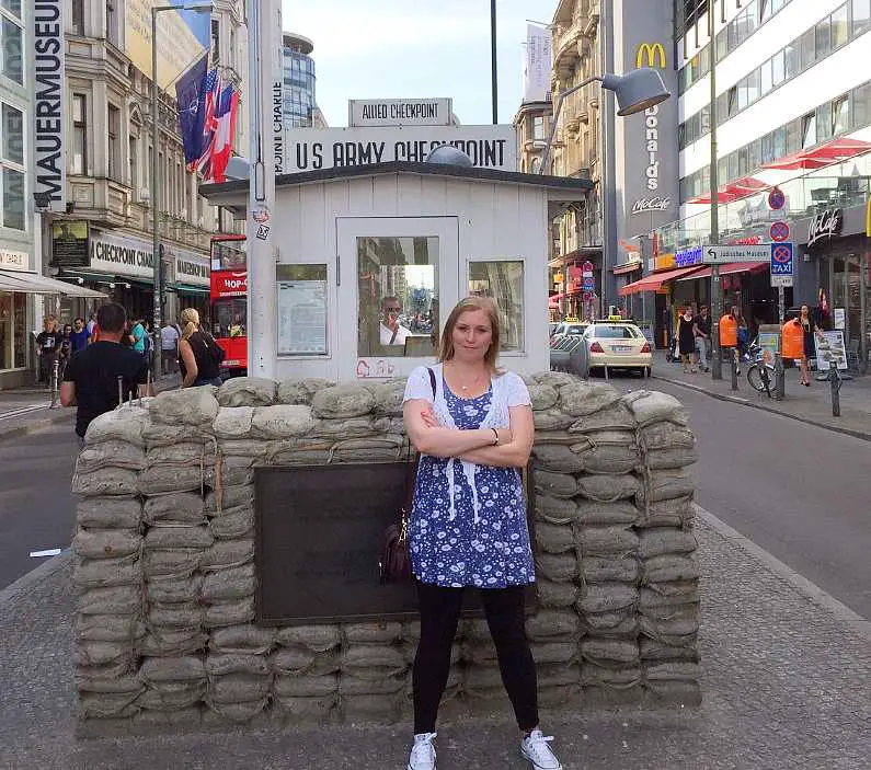 Mel stood with her arms crossed in front of Check Point Charlie, a white wooden shed check point with fake sandbags in front of it