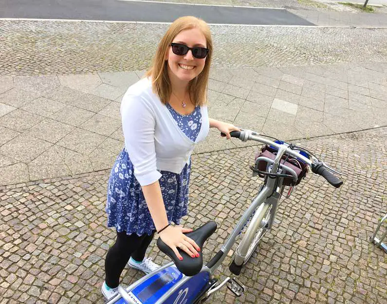 Mel holding a bike up wearing sunglasses and a blue and white dress