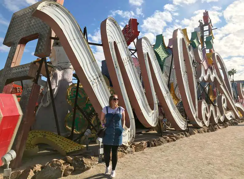 Mel wearing a jean dress and black leggings stood in front of a broken pink neon sign at the Neon Sign Museum 