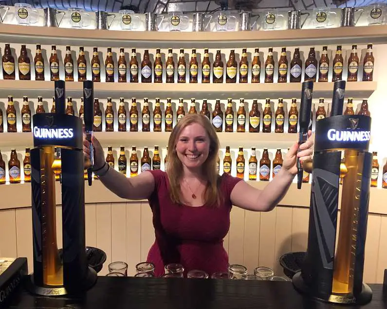 Mel stood smiling behind the Guinness Academy bar holding onto Guinness taps with rows of Guinness beer bottles behind her