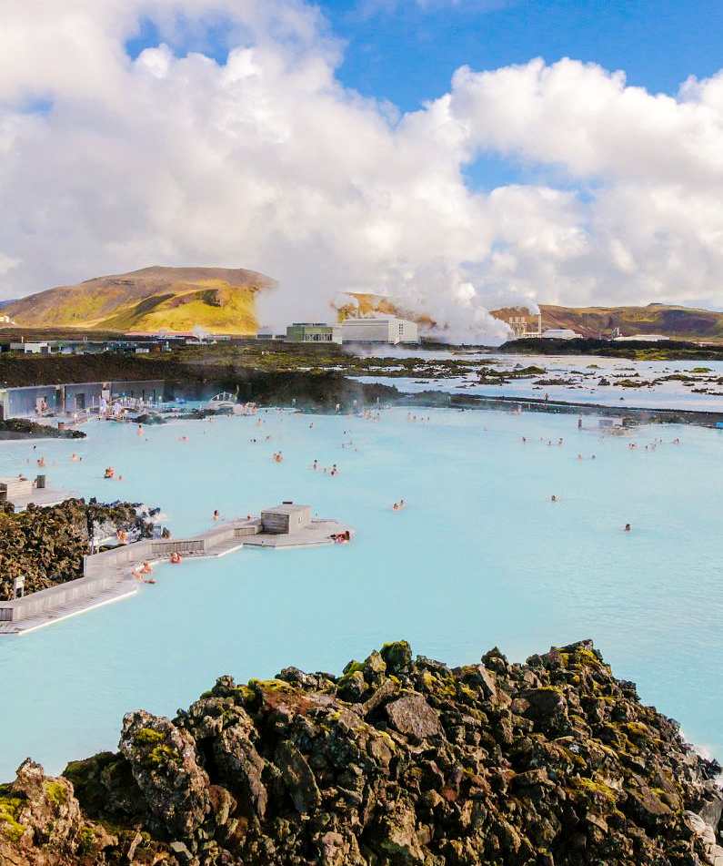 Wide view of the Blue Lagoon complex with people swimming and white smoke in the background coming from the power plant