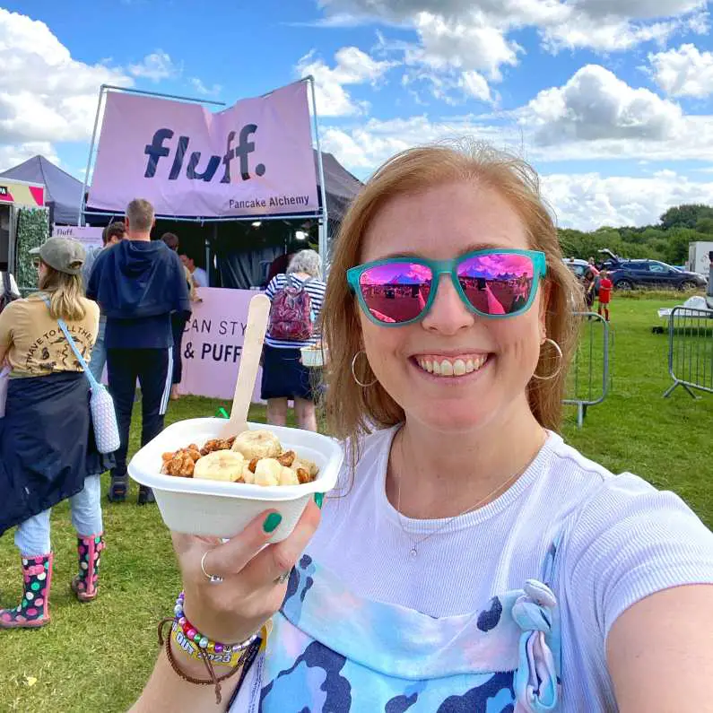 Mel holds up a dish of mini pancakes with banana slices and peanut butter sauce with the "Fluff" food stand in the background wearing blue and green overalls and pink and green glasses