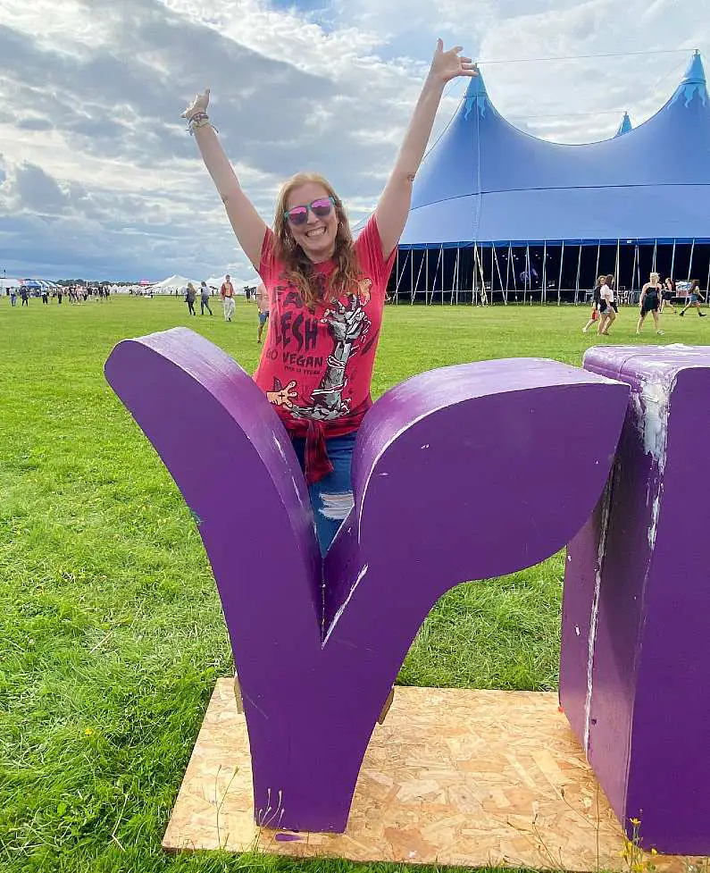 Mel holding her arms up wearing pink and green sunglasses and a red vegan t-shirt stood behind the "V" on the vegan sign at Vegan Camp Out