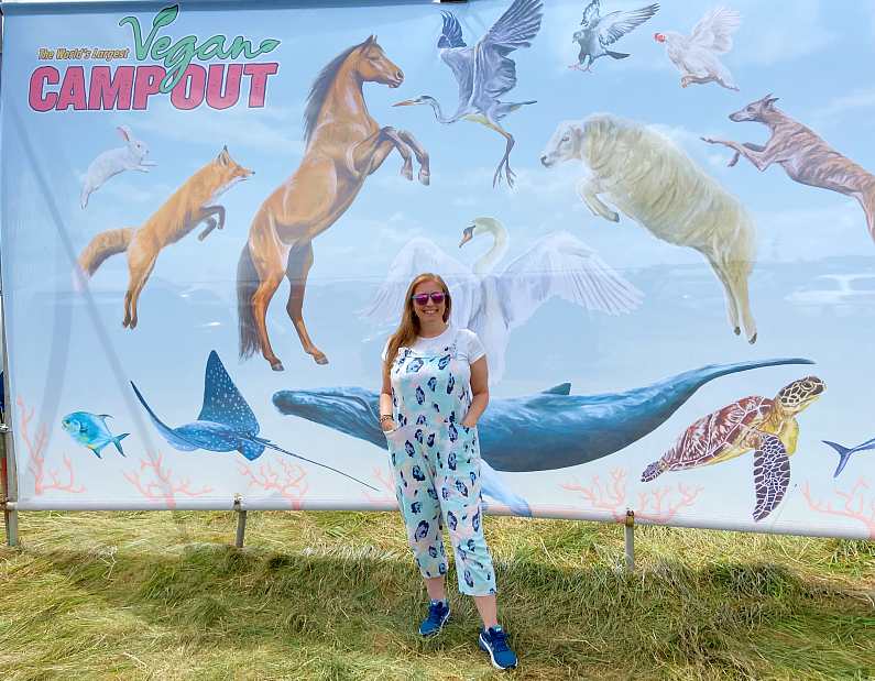 Mel stood in front of a Vegan Camp Out backdrop featuring the festival logo, farm animals and sea life wearing green and blue overalls, blue trainers and pink and green sunglasses