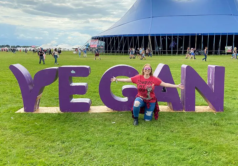 Mel knelt down in front of the "Vegan" sign at Vegan Camp Out with her arms out wearing pink and green sunglasses and a red vegan t-shirt
