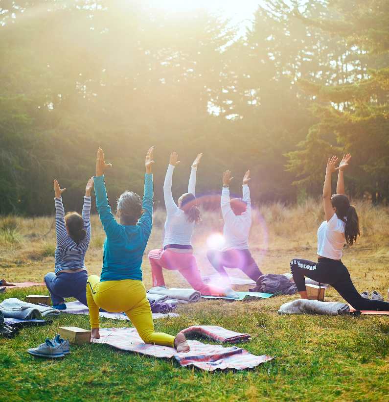Group of women in sun warrior pose doing yoga on mats outside in a forest setting