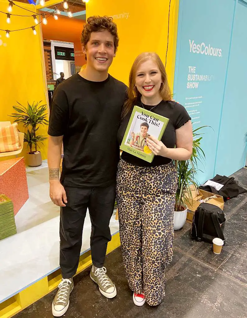 Mel wearing a black t-shirt and leopard print pants posing with Max La Manna wearing all black with green converse, smiling holding his cook book: "You can cook this!"
