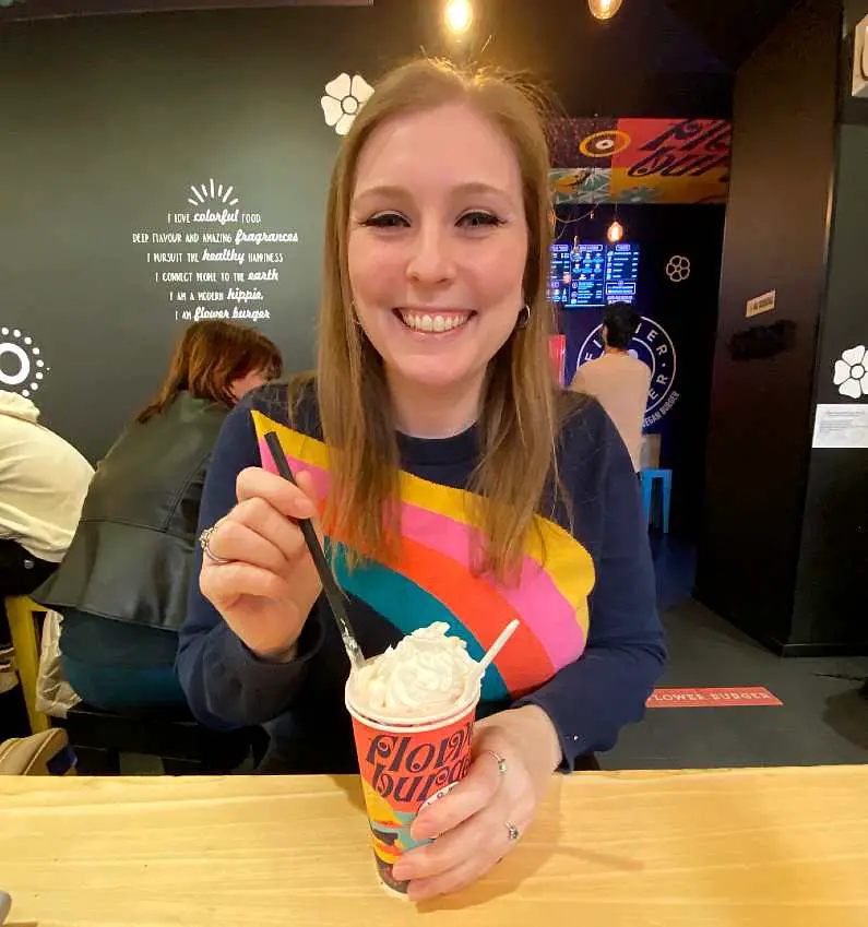 Mel smiling wearing a rainbow jumper, about to drink from a vegan milkshake with cream on top 