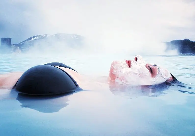 Woman up close floating on her back in the Blue Lagoon wearing a black bikini top and a white face mask