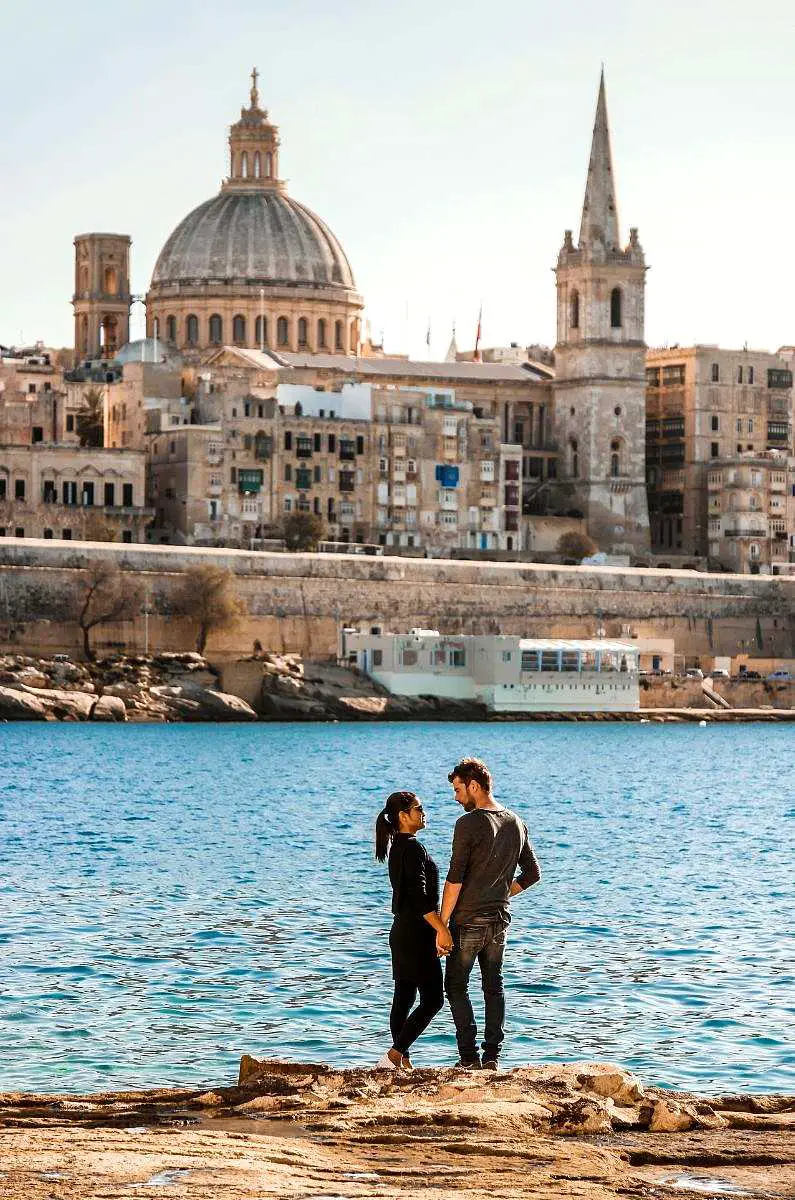 Couple standing in front of the Malta skyline holding hands and looking at each other lovingly wearing dark clothes