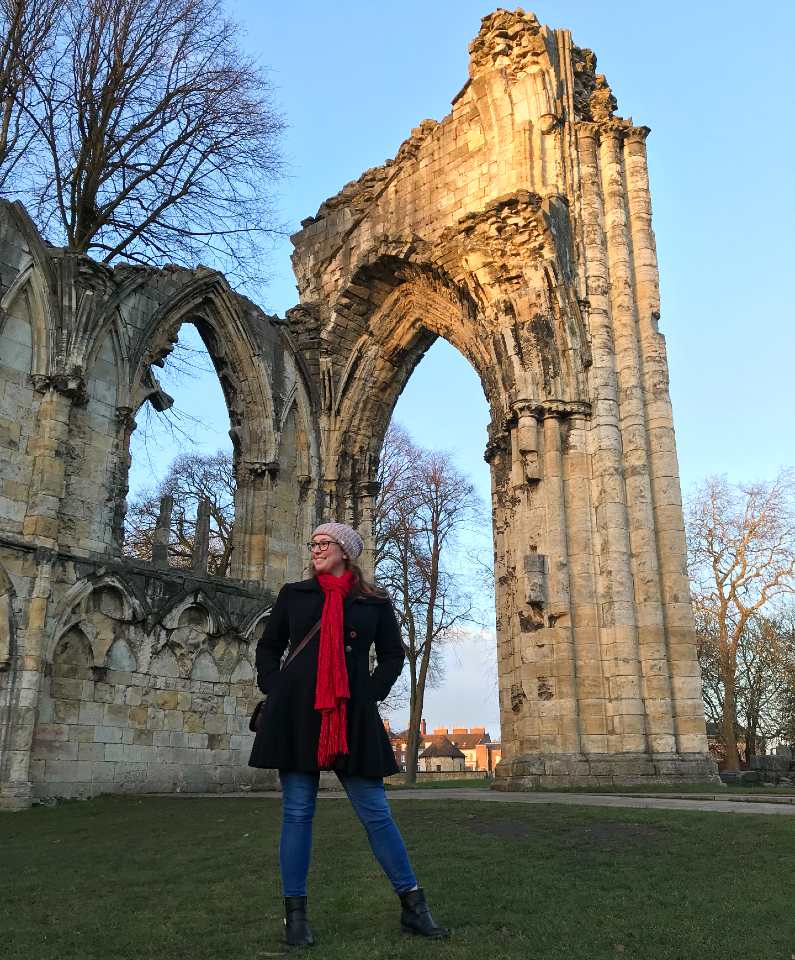 Mel stood outside Cathedral ruins in York wearing a black coat, red scraf and pink wooly hat
