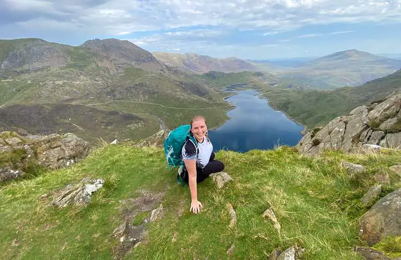 Mel smilig sat on the edge of a grassy ledge wearing a white t-shirt and green backpack just off the Watkin Path with the view of the Pyg Track on the other side of Snowdon with green peaks and a big blue lake in the background