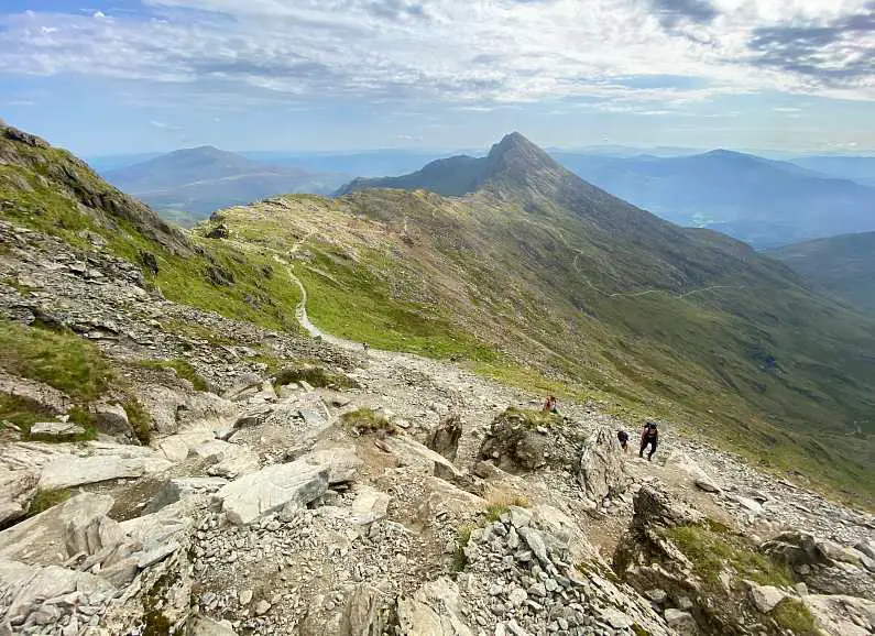 View looking down from the final ascent, a rocky patch of path that hikers are scrambling over with the Snowdonia green peaks in the background