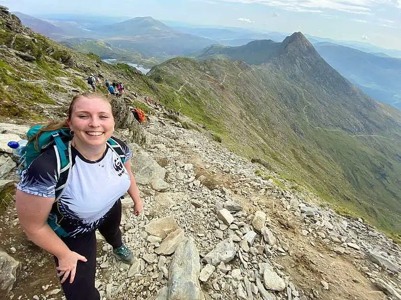 Mel looking really tired but smiling near the top of the Watkin Path final ascent with lots of scrambling rocks along the path and the green peaks of Snowdon in the background and the winding path down the mountain