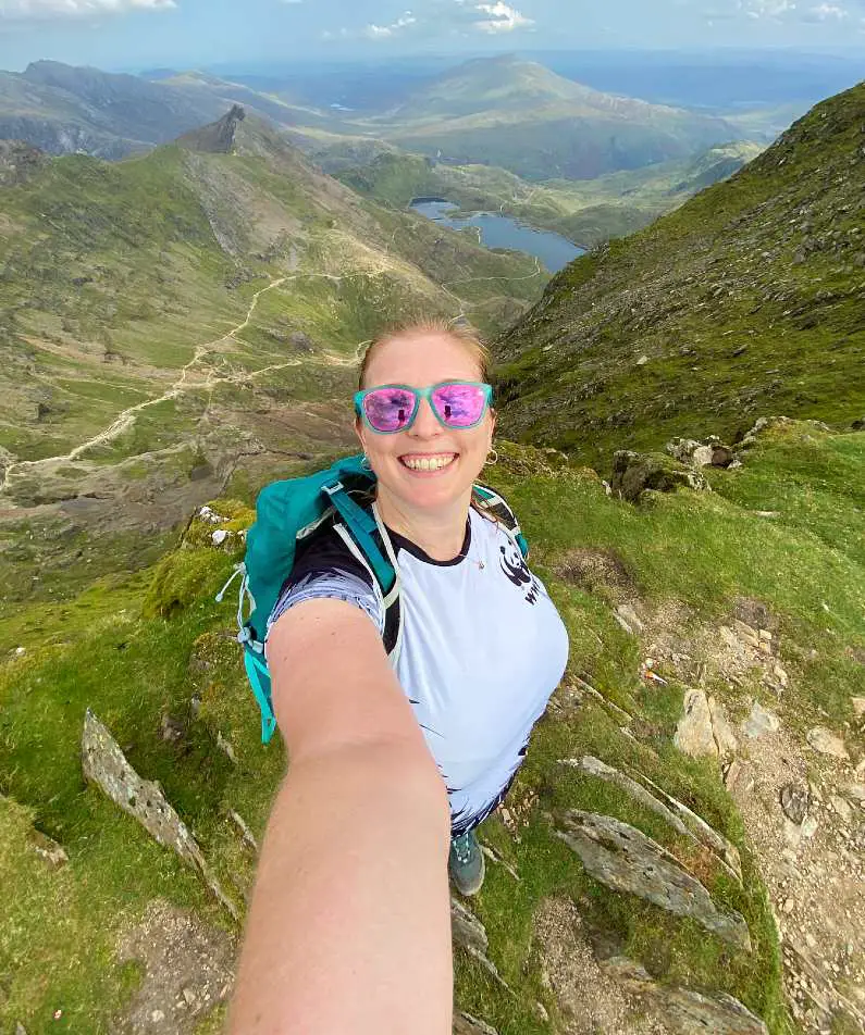 Mel taking a selfie wearing a white t-shirt, green backpack and pink sunglasses with the green peaks and blue lakes of Snowdonia National Park in the background from the summit