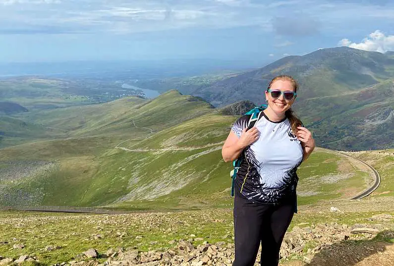Mel wearing a white t-shirt, black leggings and a green backpack stood at the top of the Llanberis Path with the path winding in the background down the mountainside with lovely green peaks in the background