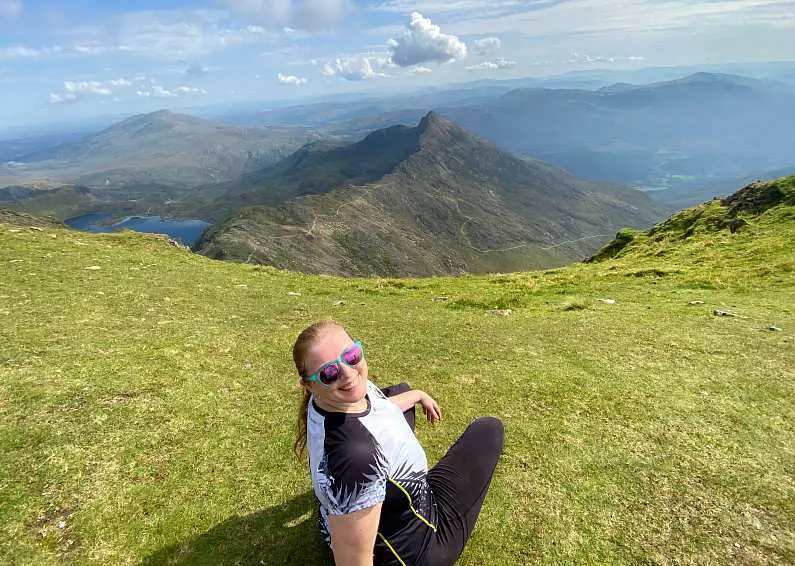 Mel wearing a white t-shirt, black leggings and pink sunglasses turning around smiling at the top of the Watkin Path with the path winding down the mountain in the background with peaks and lakes