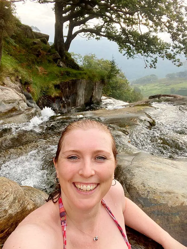 Mel taking a selfie swimming in the Watkin Path Waterfall pool wearing a red bikini with the trees and hills of Snowdon in the background